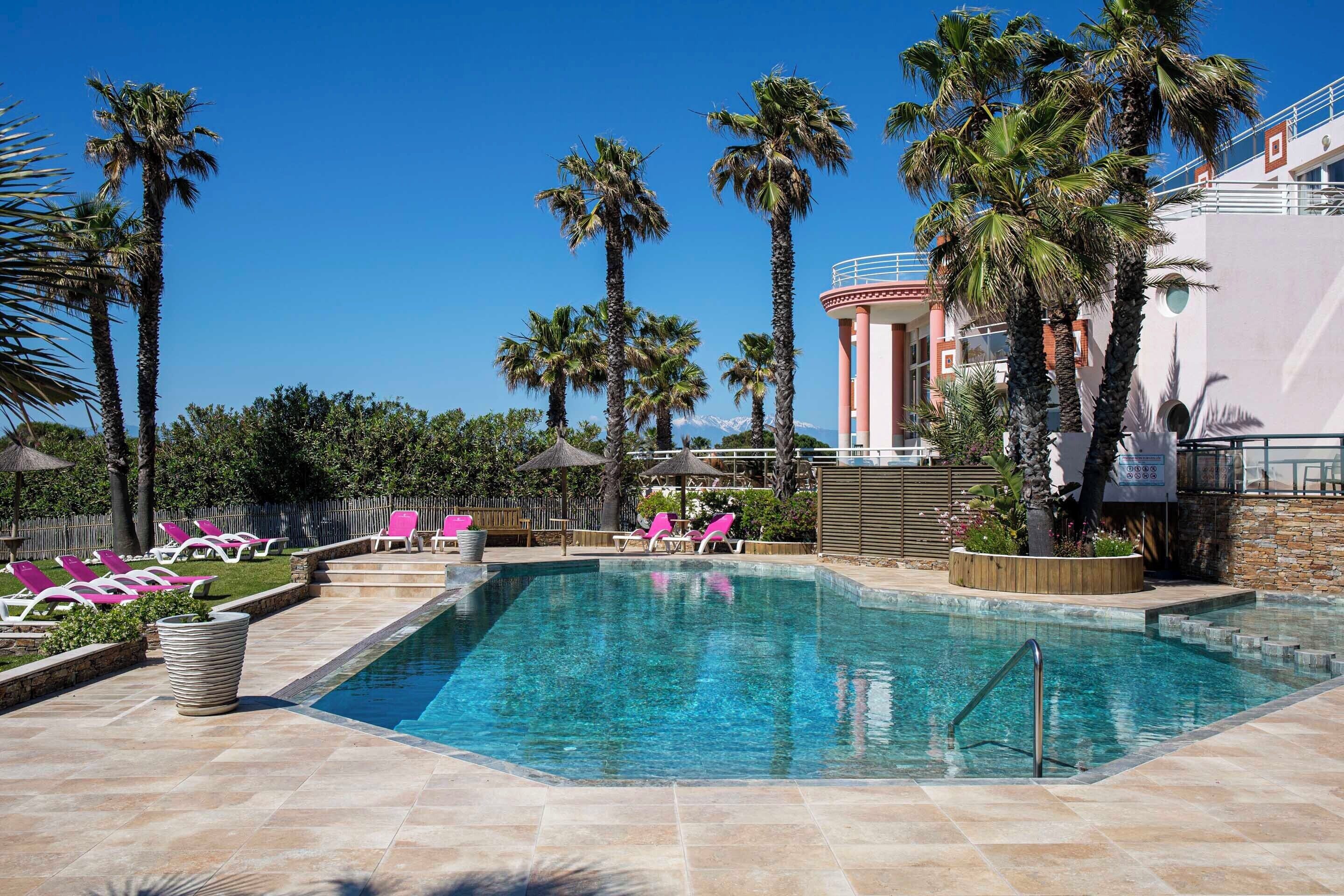 A swimming pool with lounge chairs, palm trees, and a building in the background.