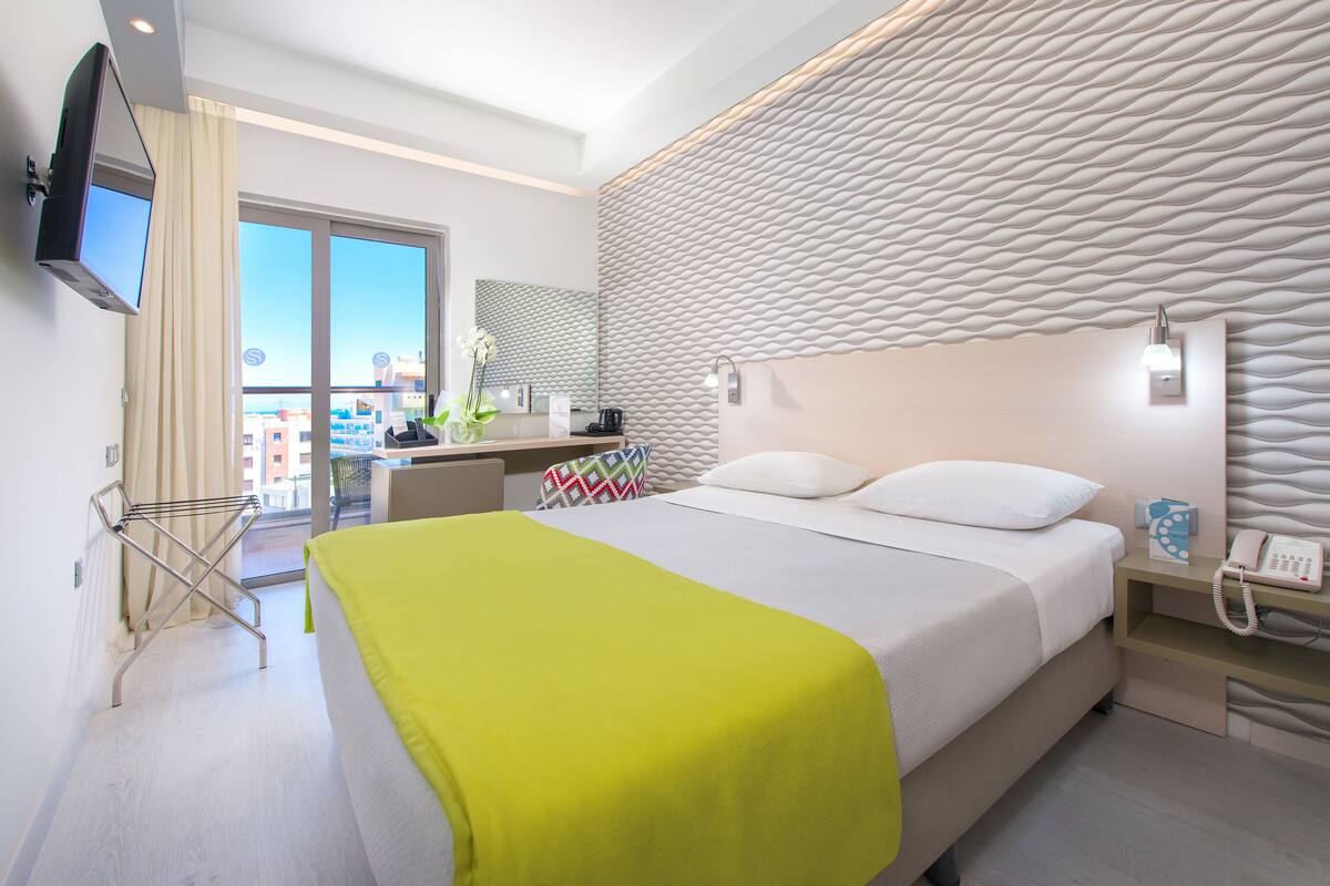 A modern hotel room with a large bed, a flat-screen TV mounted on the wall, a bedside table with a lamp, and a view of the city through the window.