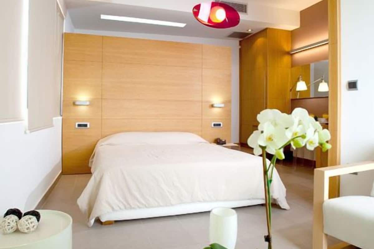 A modern hotel room with a large bed, a bedside table with a vase of flowers, a glass coffee table, and a wooden headboard.