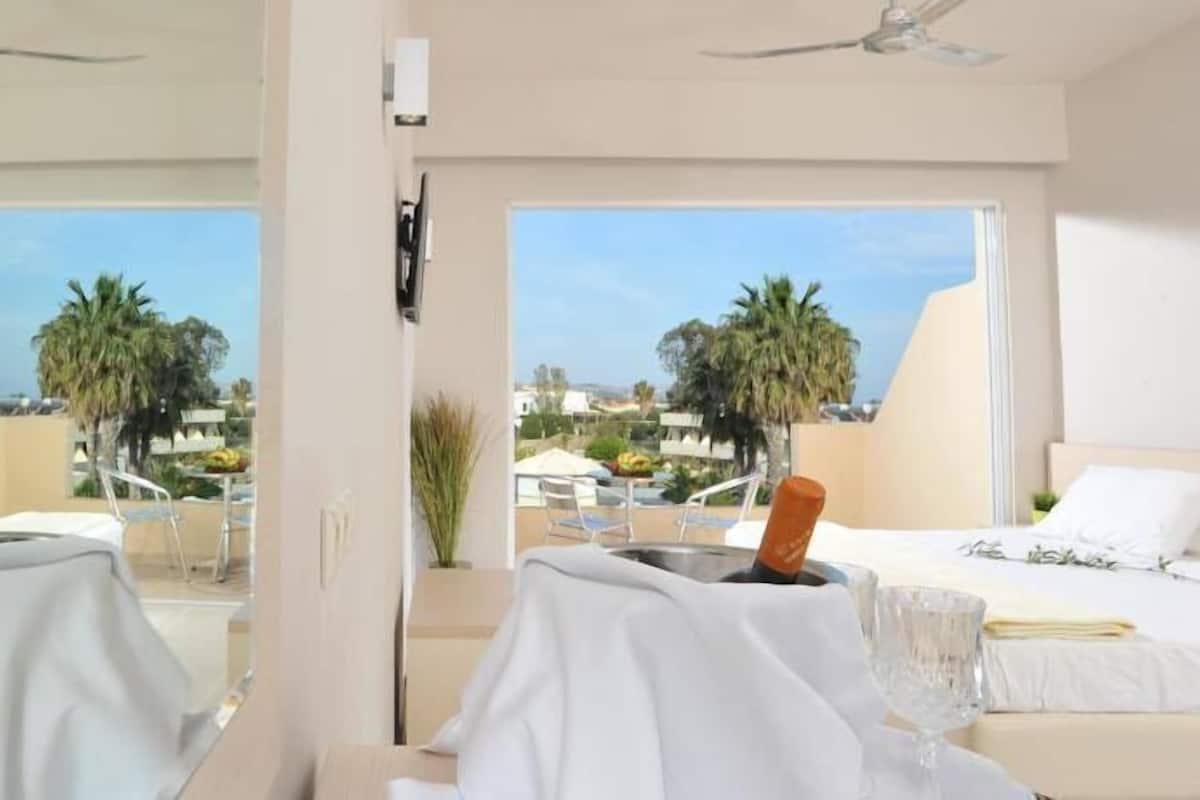 A hotel room with a bed, a table with a glass, and a view of palm trees and buildings.