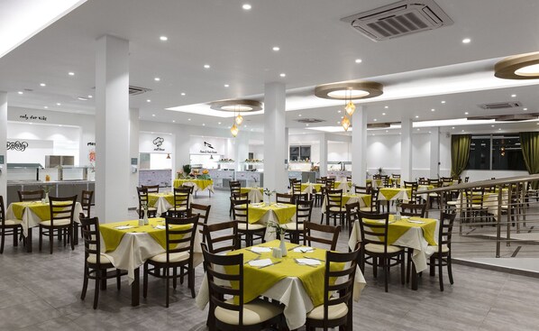 A spacious restaurant with multiple dining tables and chairs, a kitchen area in the background, and a staircase leading to an upper level. - Olive Garden Hotel (Rhodes)