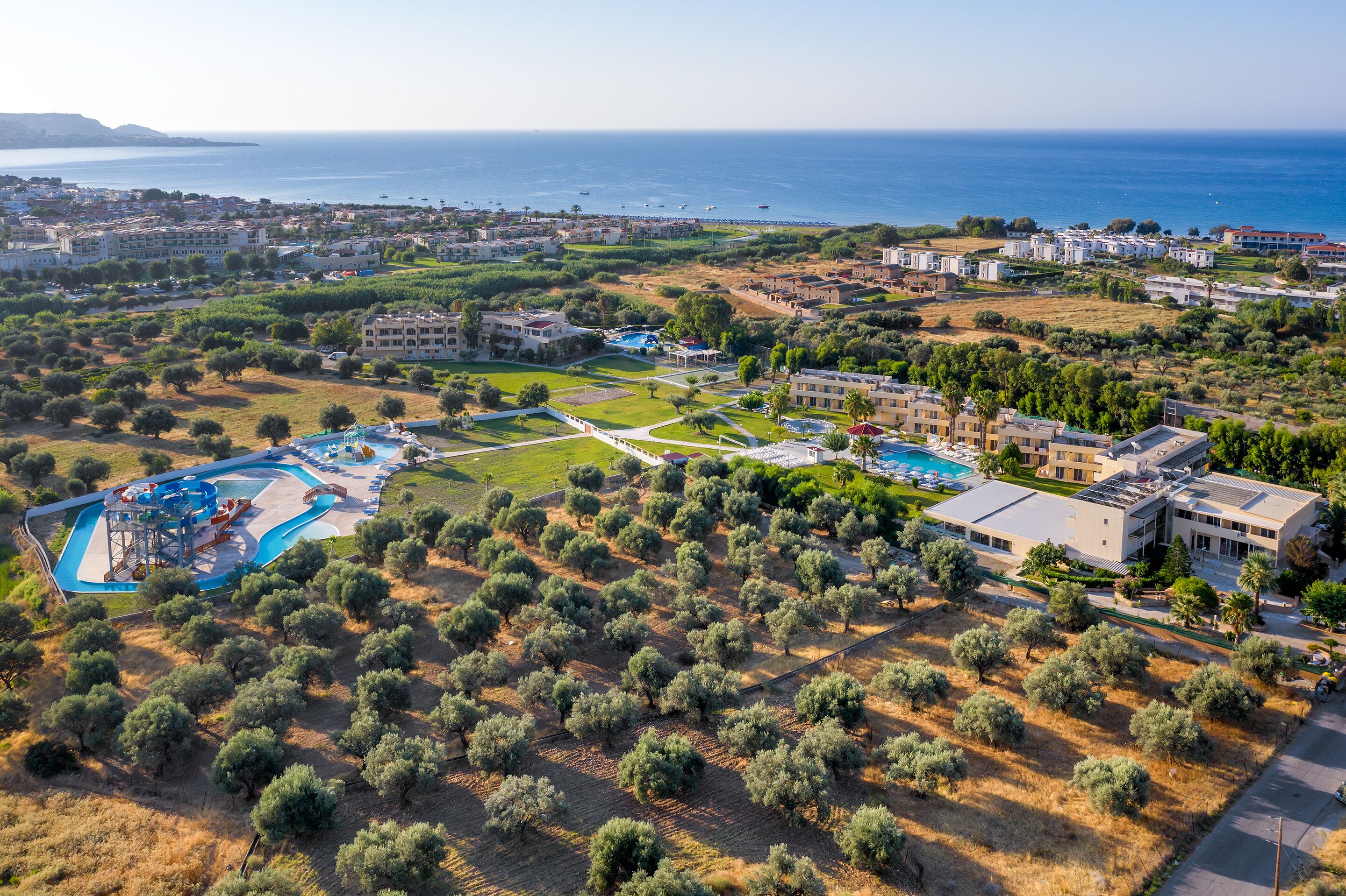An aerial view of a coastal resort with multiple buildings, a swimming pool, and a landscaped area with olive trees.