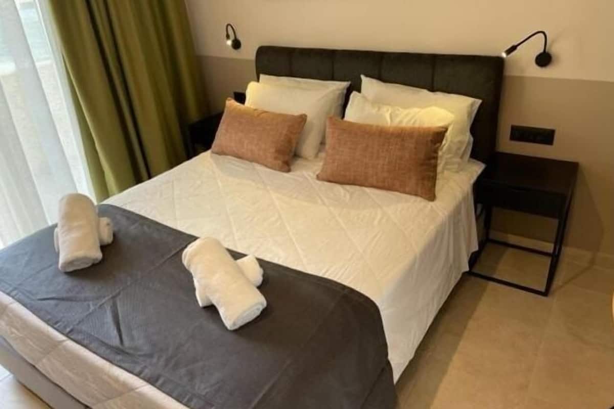 A neatly made bed with a dark headboard, white bedding, and two pillows. There are two bedside tables, one with a lamp and the other with a power outlet.