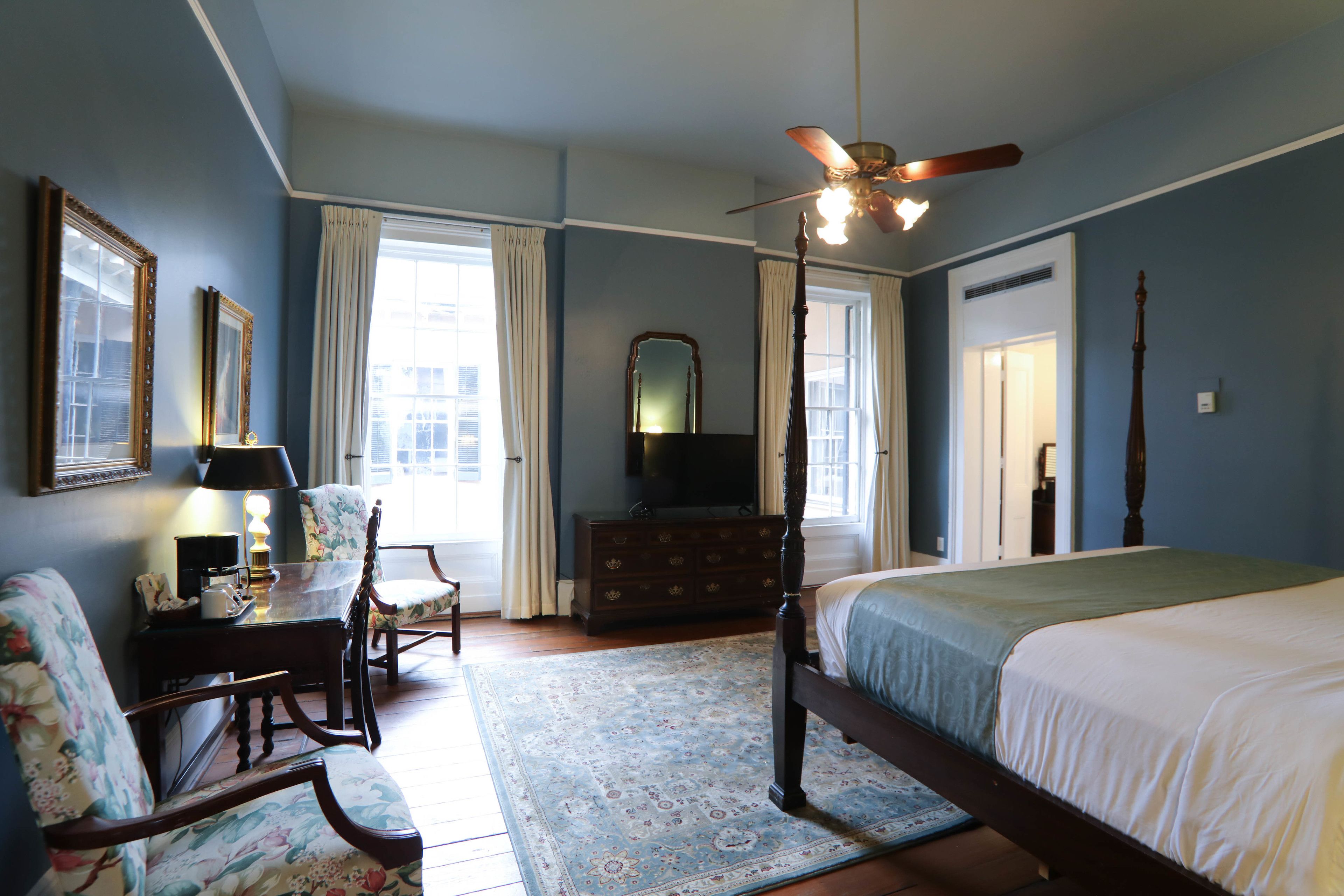 A bedroom with a four-poster bed, a ceiling fan, a dresser, a mirror, and a desk with a lamp.