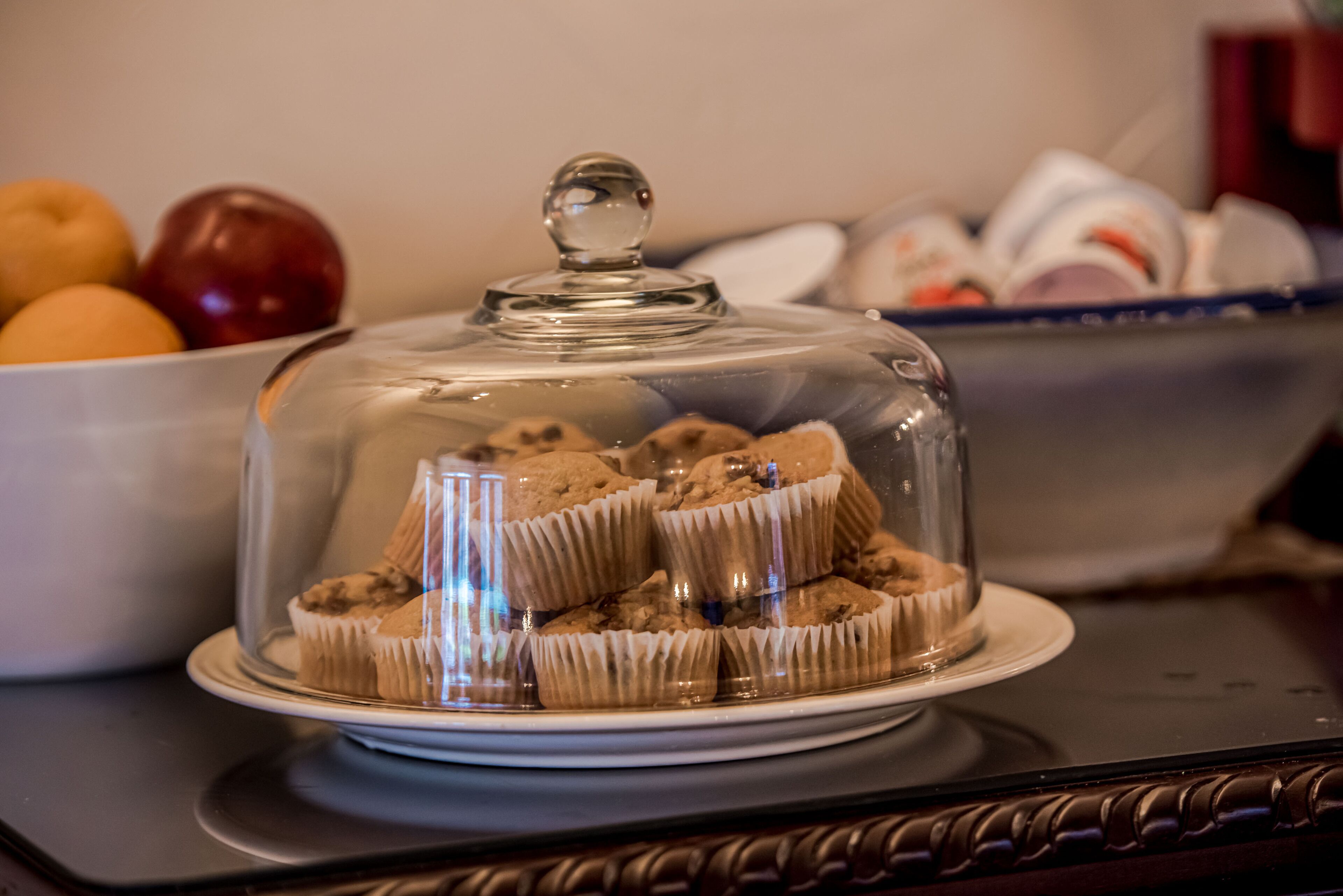 A glass-covered dessert stand with cupcakes on a white plate, a bowl of fruits in the background, and a blue bowl with items.