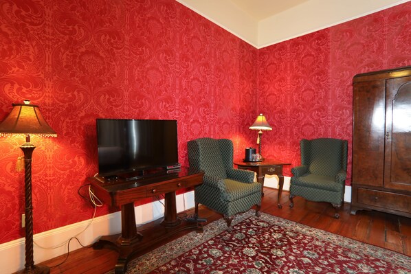 A room with a red patterned wallpaper, a television, two armchairs, a wooden cabinet, and a patterned rug.