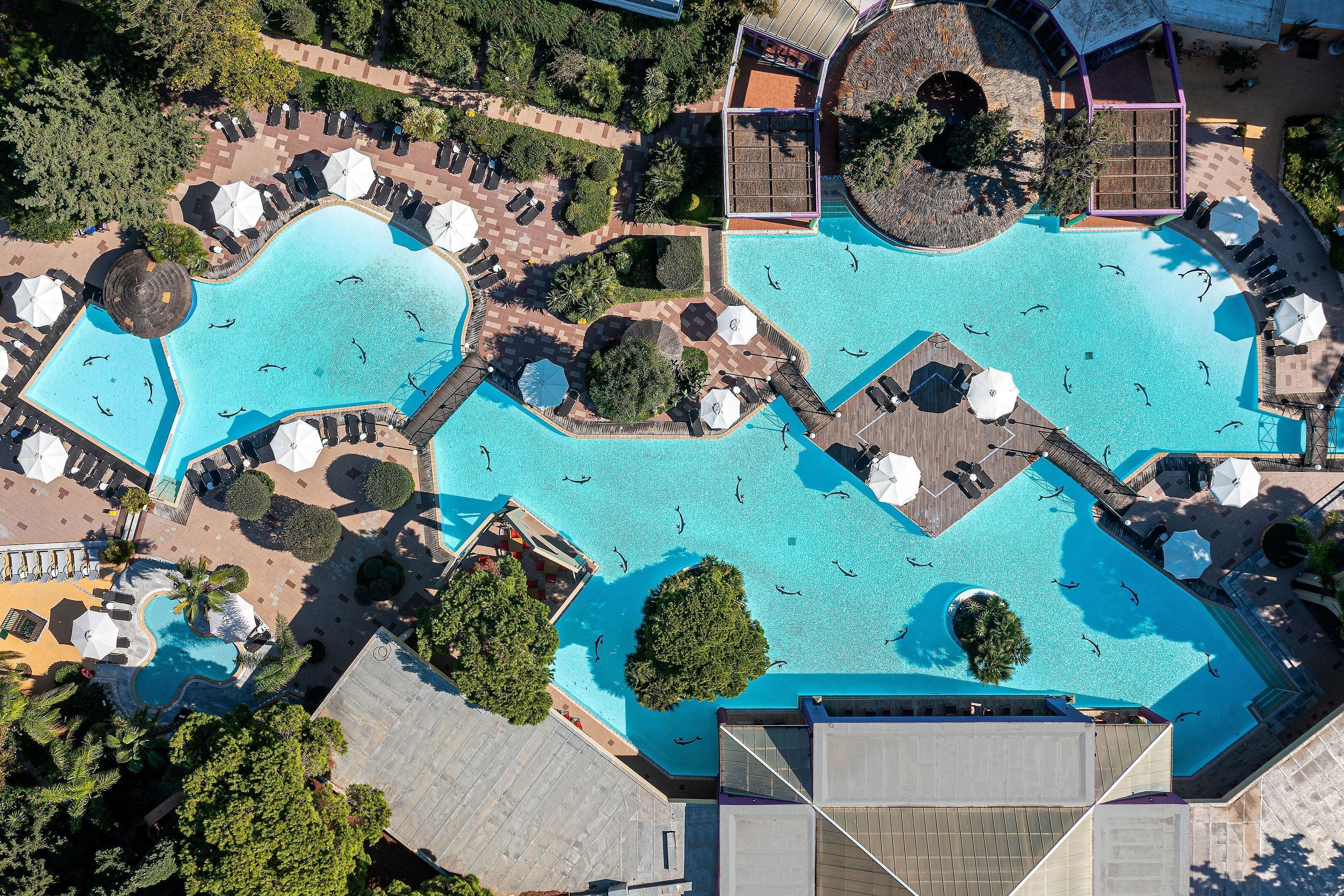 Vista aérea de un hotel con varias piscinas, tumbonas y zonas a la sombra.