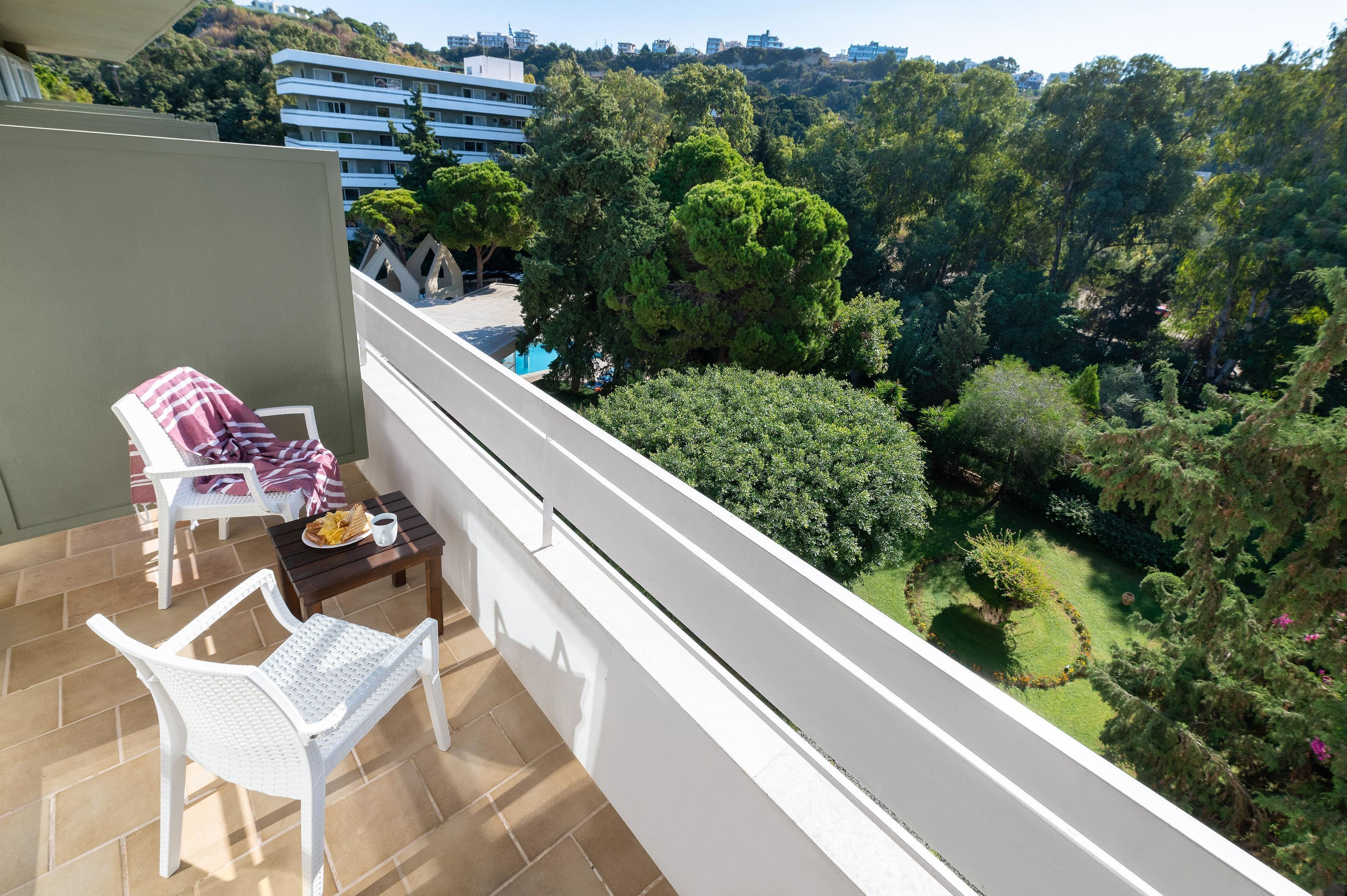 A balcony with a white chair, a small table with a plate of food, and a view of a lush green landscape with trees and a building in the background.