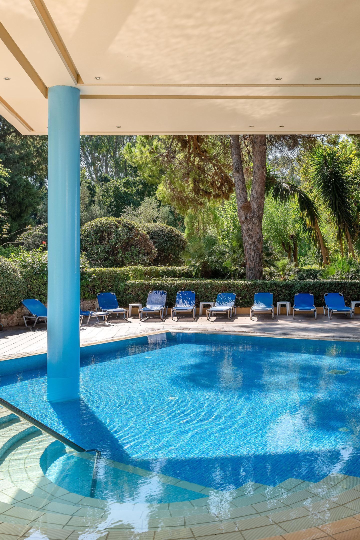 Ein Swimmingpool mit Liegestühlen unter einer Überdachung, umgeben von Grünflächen.