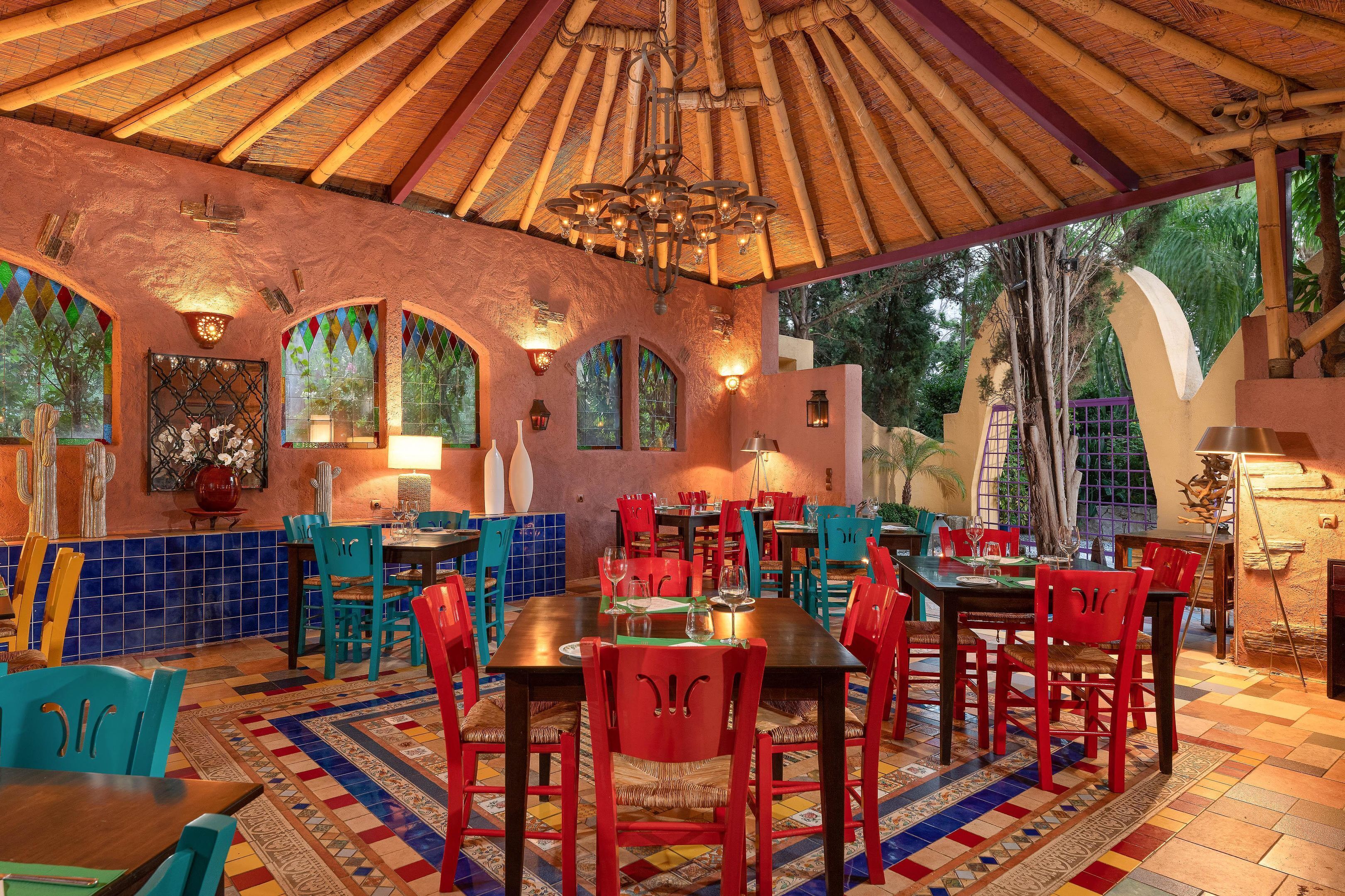 A restaurant with a rustic interior, featuring a thatched roof, colorful tiled walls, and a variety of seating arrangements including tables and chairs.