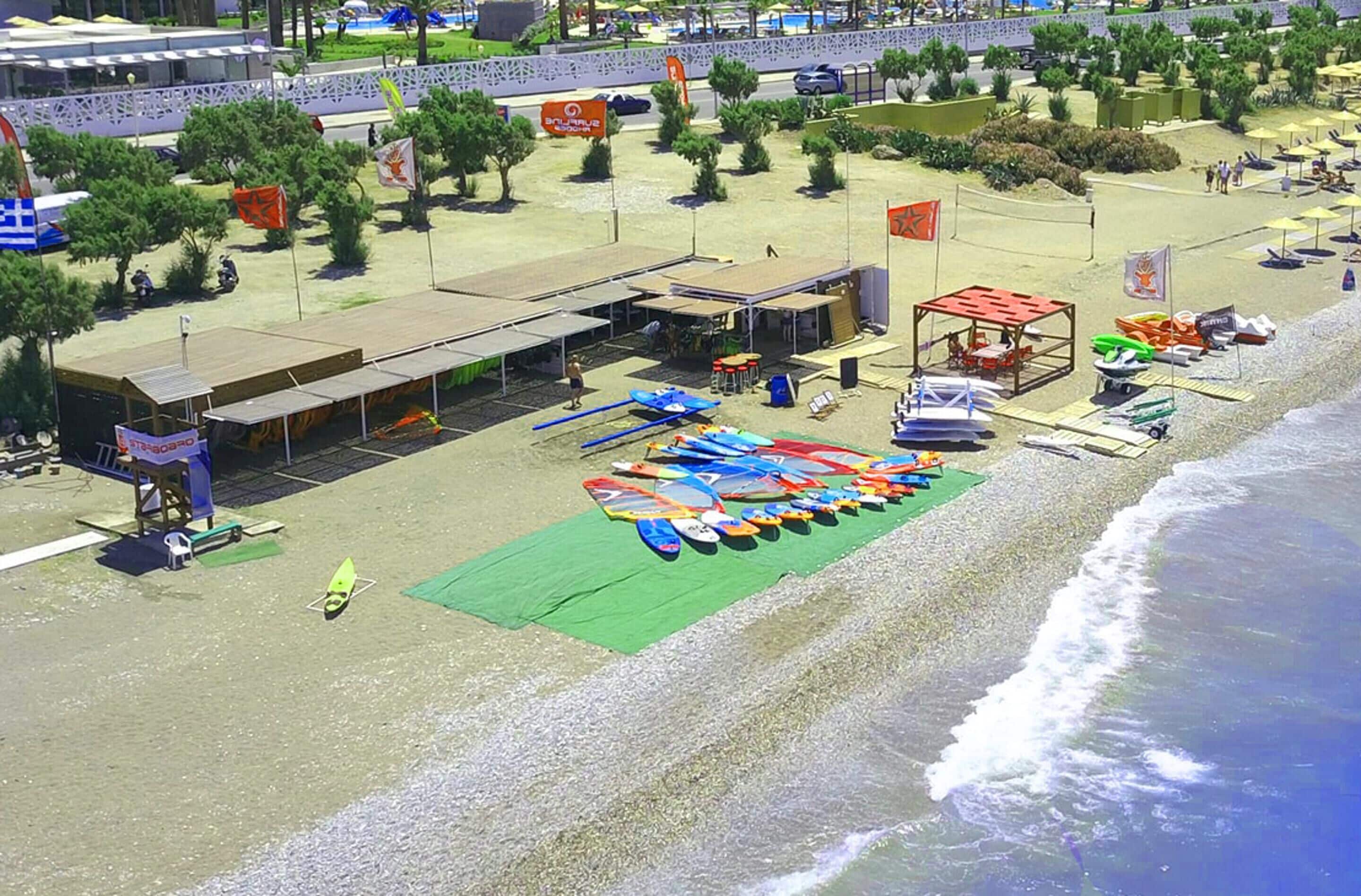 An aerial view of a beachside area with a building, a row of colorful kayaks, and a sandy beach.