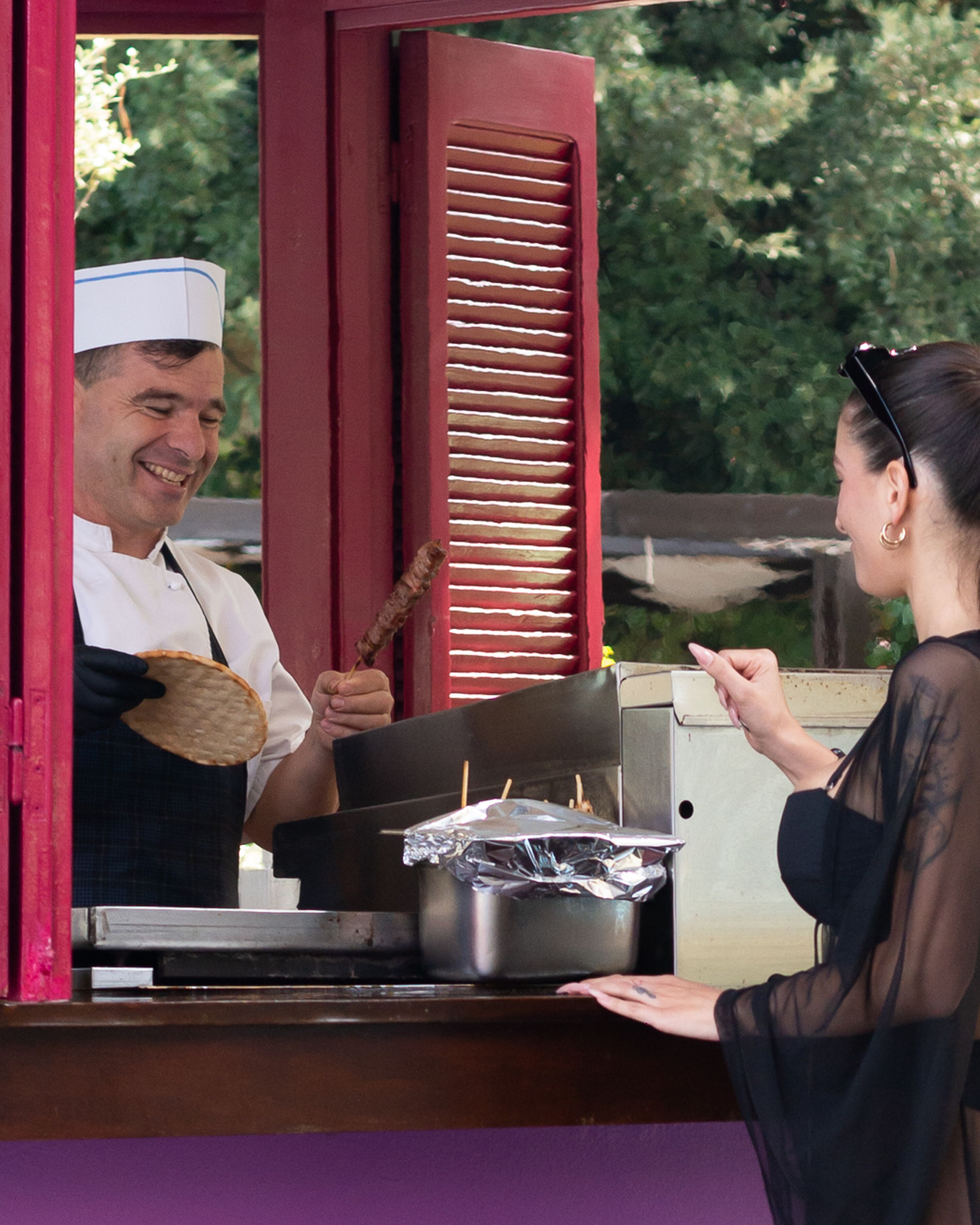 Un vendeur de nourriture avec un gril et des ustensiles de cuisine, un client et une fenêtre à cadre rouge.