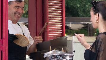 Un vendedor de comida con una parrilla y utensilios de cocina, un cliente y un marco de ventana rojo.