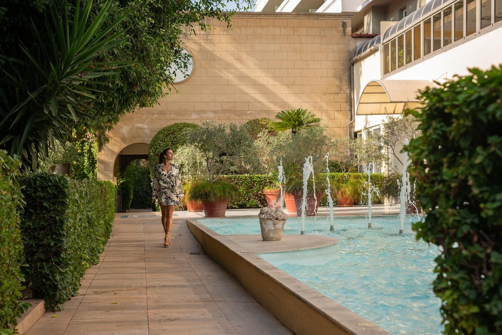 A woman walking along a tiled pathway beside a pool with water fountains, surrounded by lush greenery and a building with a large arched window.