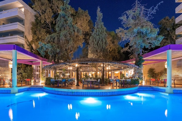 A pool area with a covered seating area, surrounded by trees and a building with balconies. - Dionysos Hotel (Rhodes)