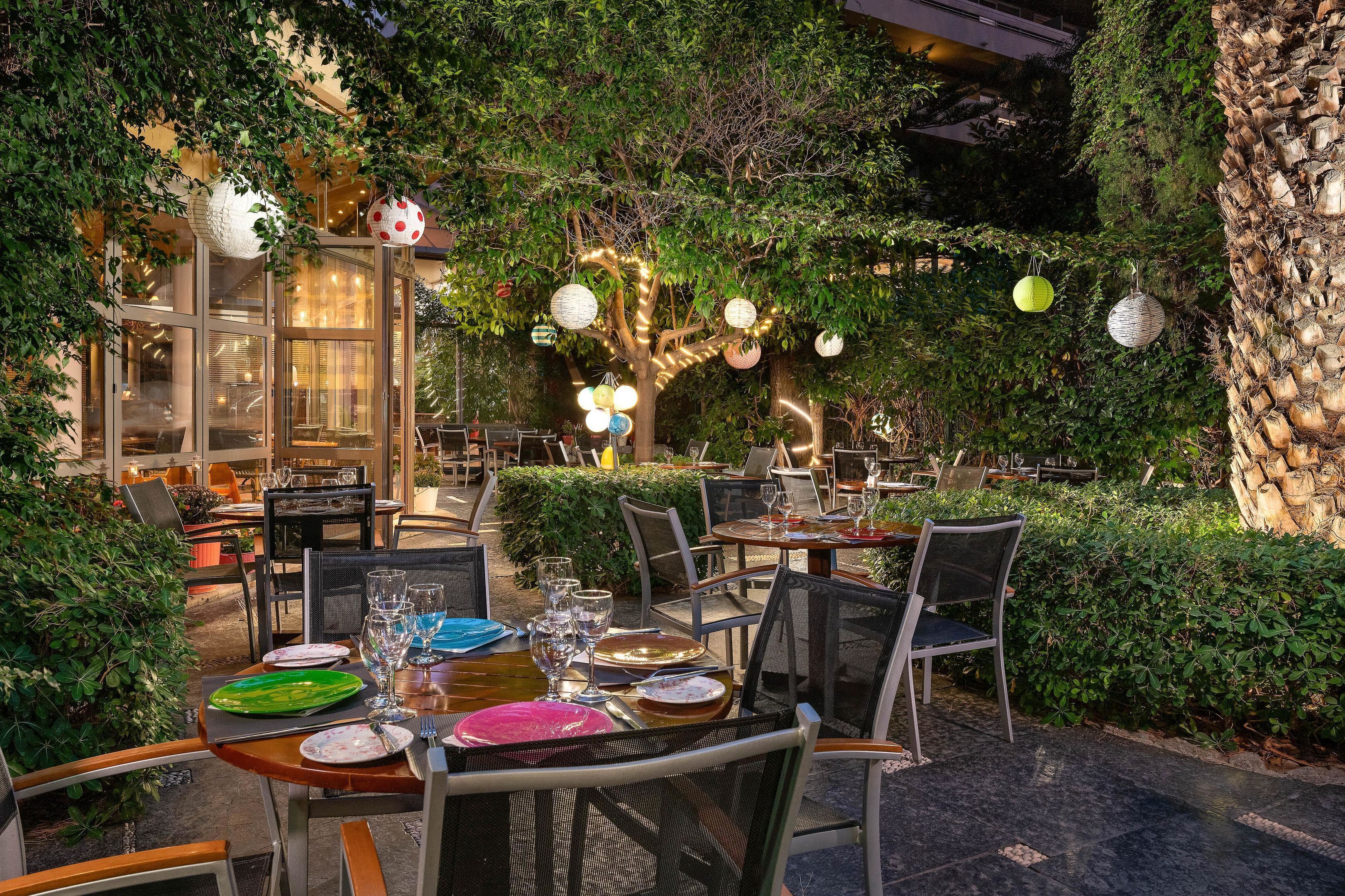 Un área de comedor al aire libre con mesas dispuestas para los comensales, rodeada de exuberante follaje e iluminación decorativa.