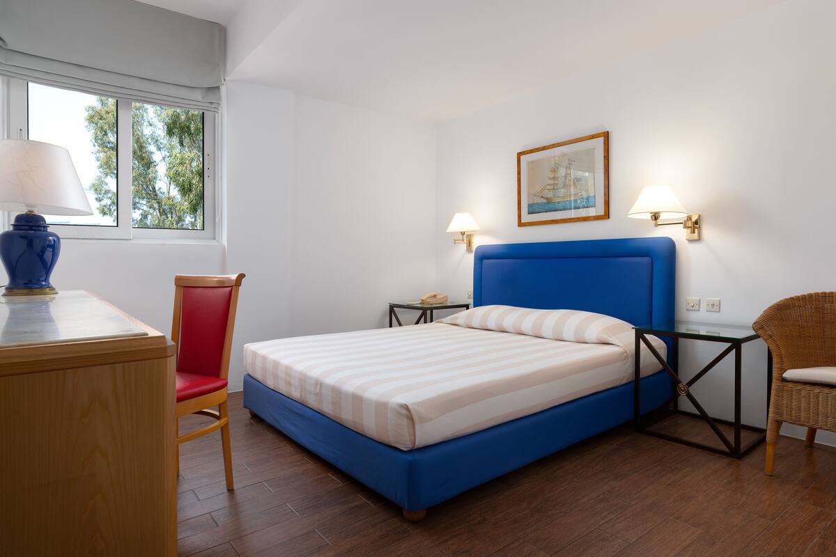 A bedroom with a bed, a desk, a chair, a nightstand, and a lamp.