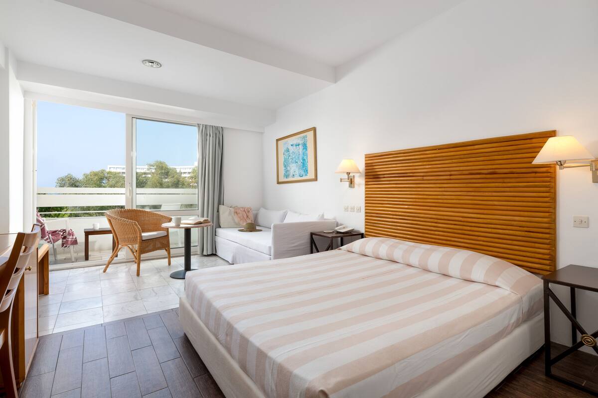 A hotel room with a large bed, a desk, a chair, a window with a view, and a wooden headboard.