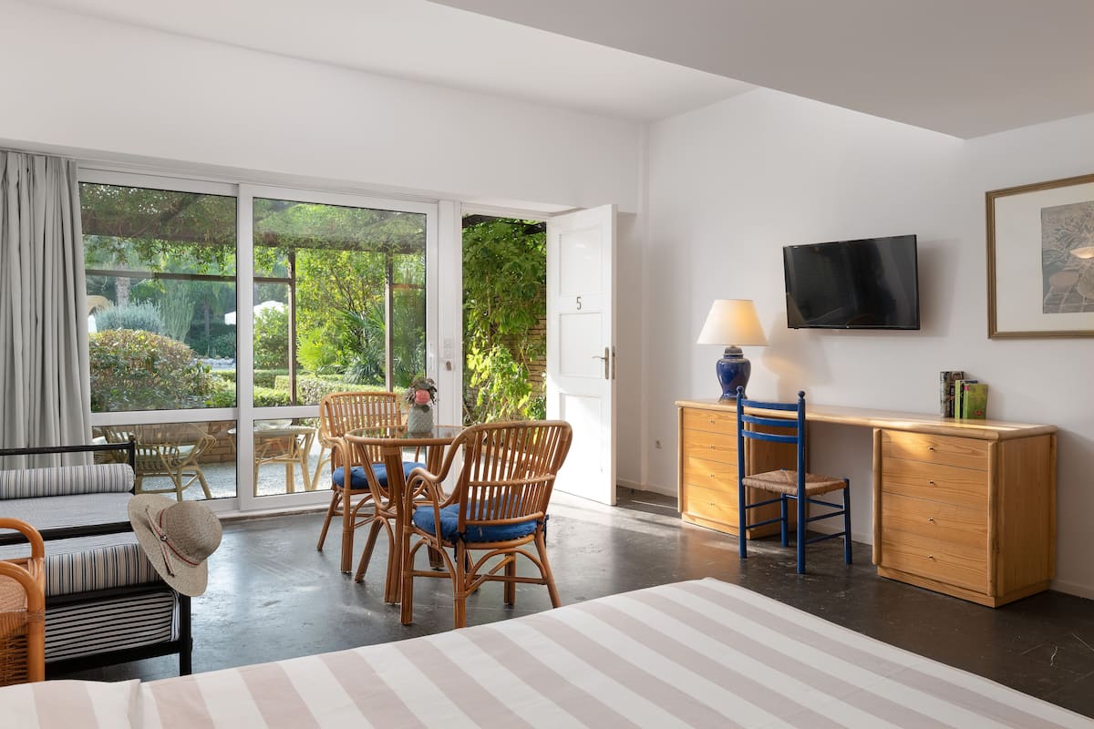 A room with a large sliding glass door leading to a garden, a wooden desk with a lamp, and a television mounted on the wall.