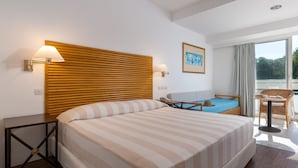 A hotel room with a large bed, a wooden headboard, two bedside lamps, a small table, a chair, and a view of the outdoors through a large window. - Dionysos Hotel (Rhodes)