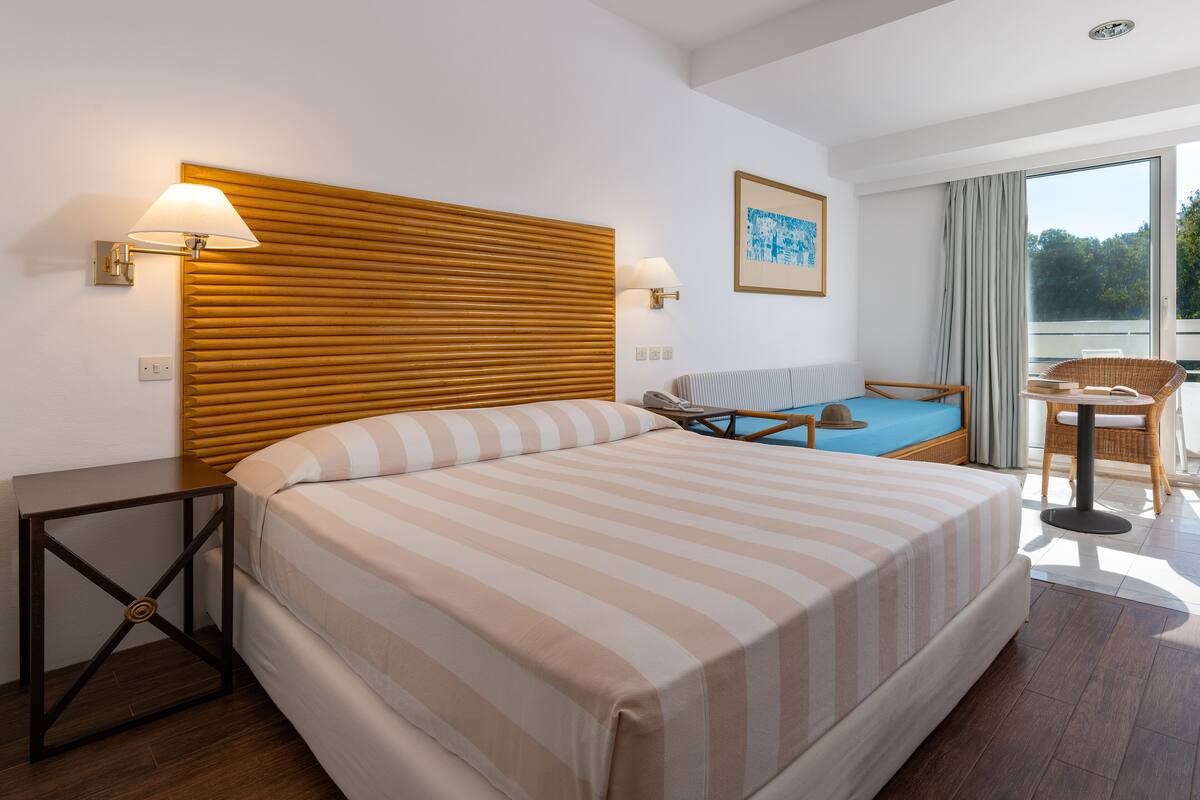 A hotel room with a large bed, a wooden headboard, two bedside lamps, a small table, a chair, and a view of the outdoors through a large window.