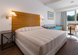 A hotel room with a large bed, a wooden headboard, two bedside lamps, a small table, a chair, and a view of the outdoors through a large window.