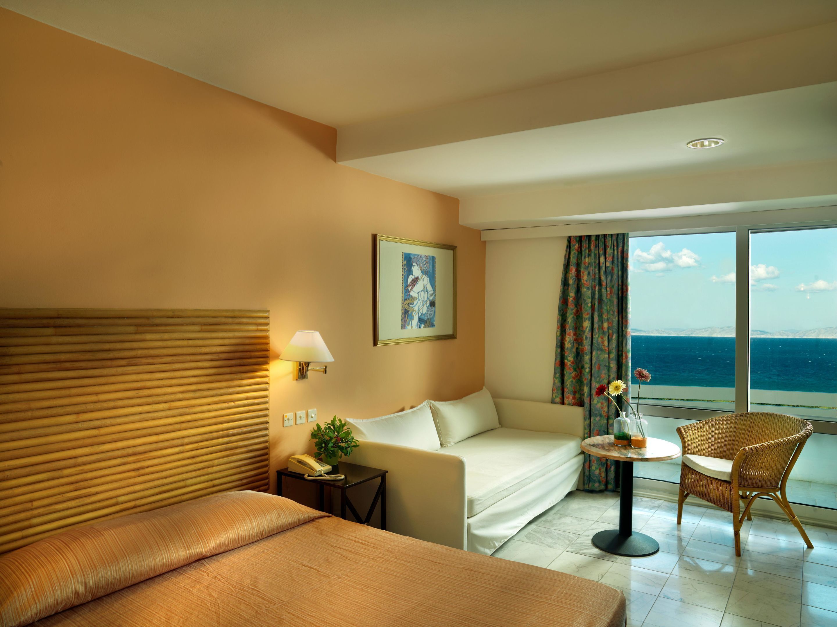 A hotel room with a bed, a sofa, a small table with a lamp, a chair, and a view of the sea.