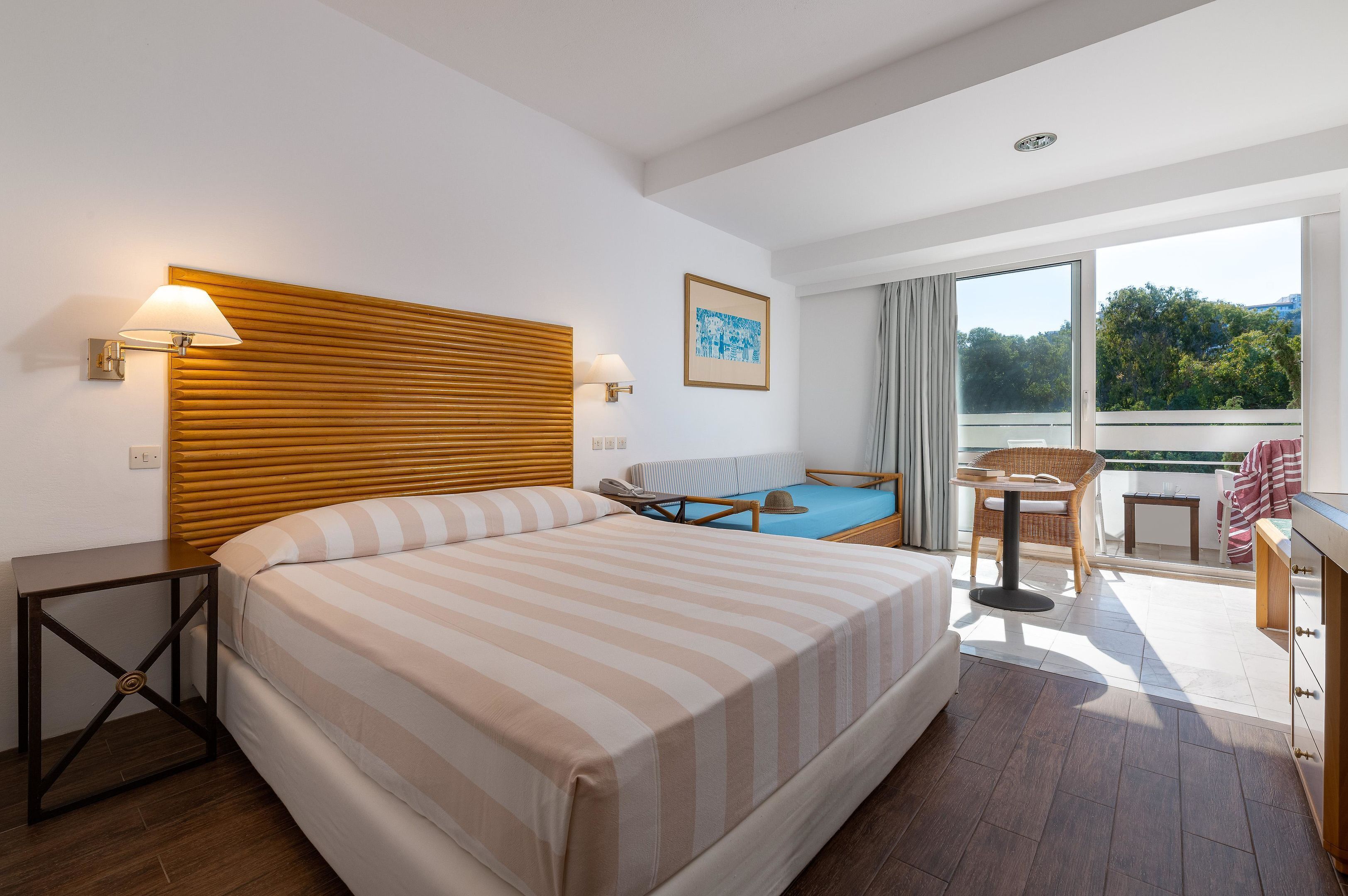 A hotel room with a large bed, a small desk, a chair, a window with a view of trees, and a balcony with a table and chairs.