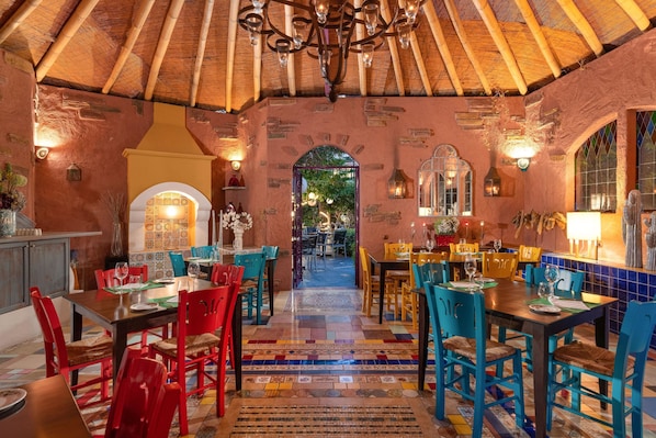 A restaurant with a rustic interior, featuring a thatched roof, colorful tiled floors, and a variety of seating arrangements. - Dionysos Hotel (Rhodes)