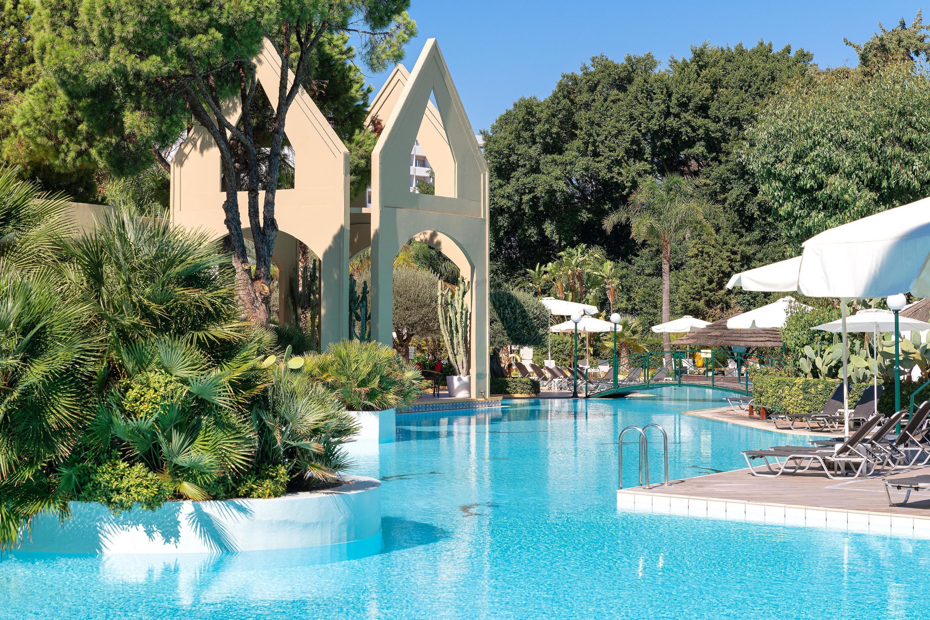 Una piscina con sillas de descanso y sombrillas, rodeada de palmeras y un edificio con un arco apuntado.