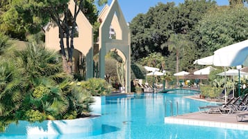 Una piscina con sillas de descanso y sombrillas, rodeada de palmeras y un edificio con un arco apuntado.