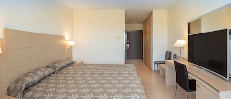 A hotel room with a bed, bedside lamps, a desk with a chair, a television, and a door leading to another room.