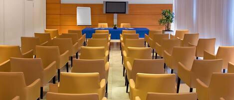 A conference room with rows of chairs, a table with a blue tablecloth, a television mounted on the wall, and a potted plant.