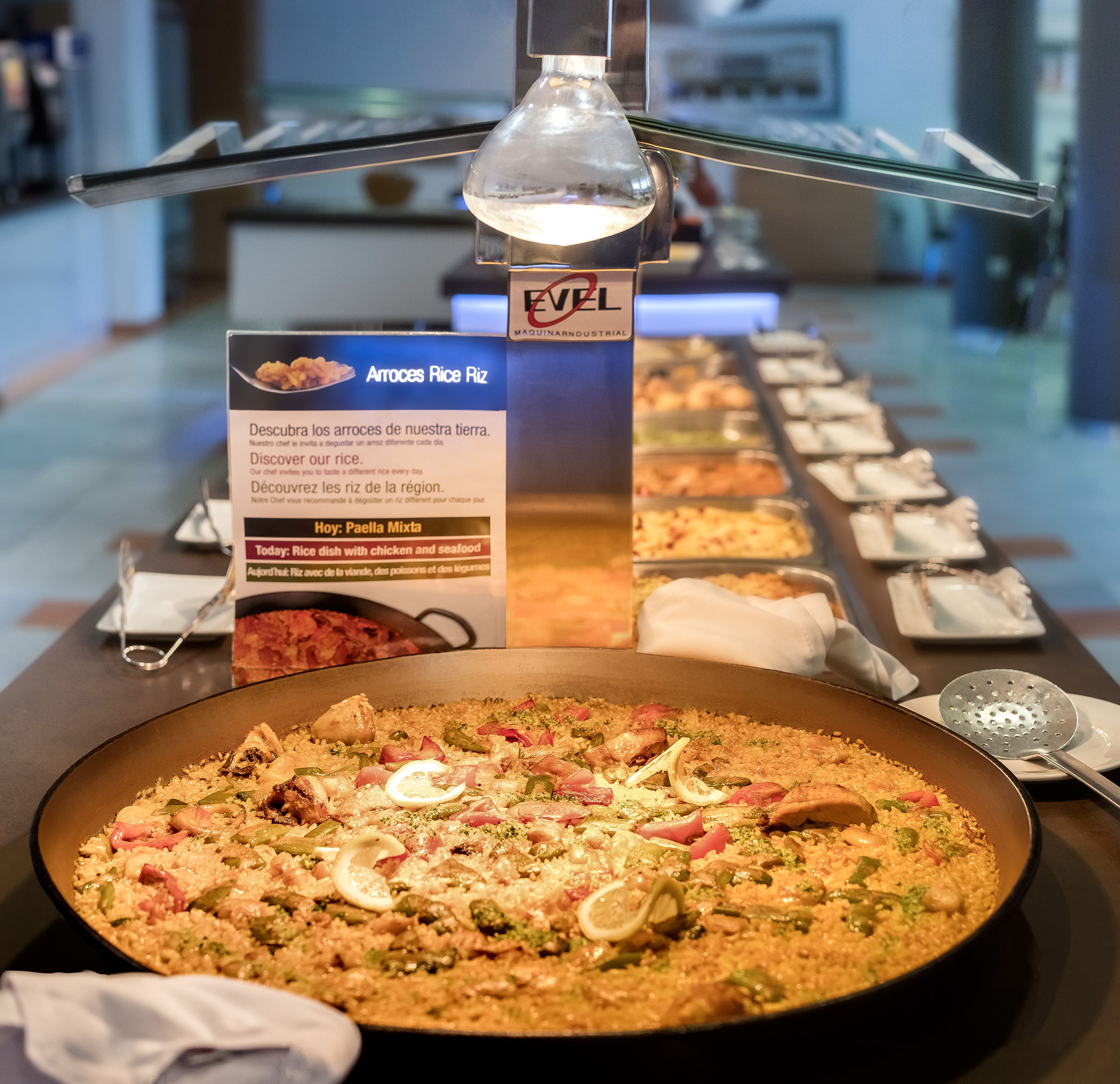 Una mesa de buffet con un plato de paella en el centro, rodeada de varios recipientes y utensilios para servir.
