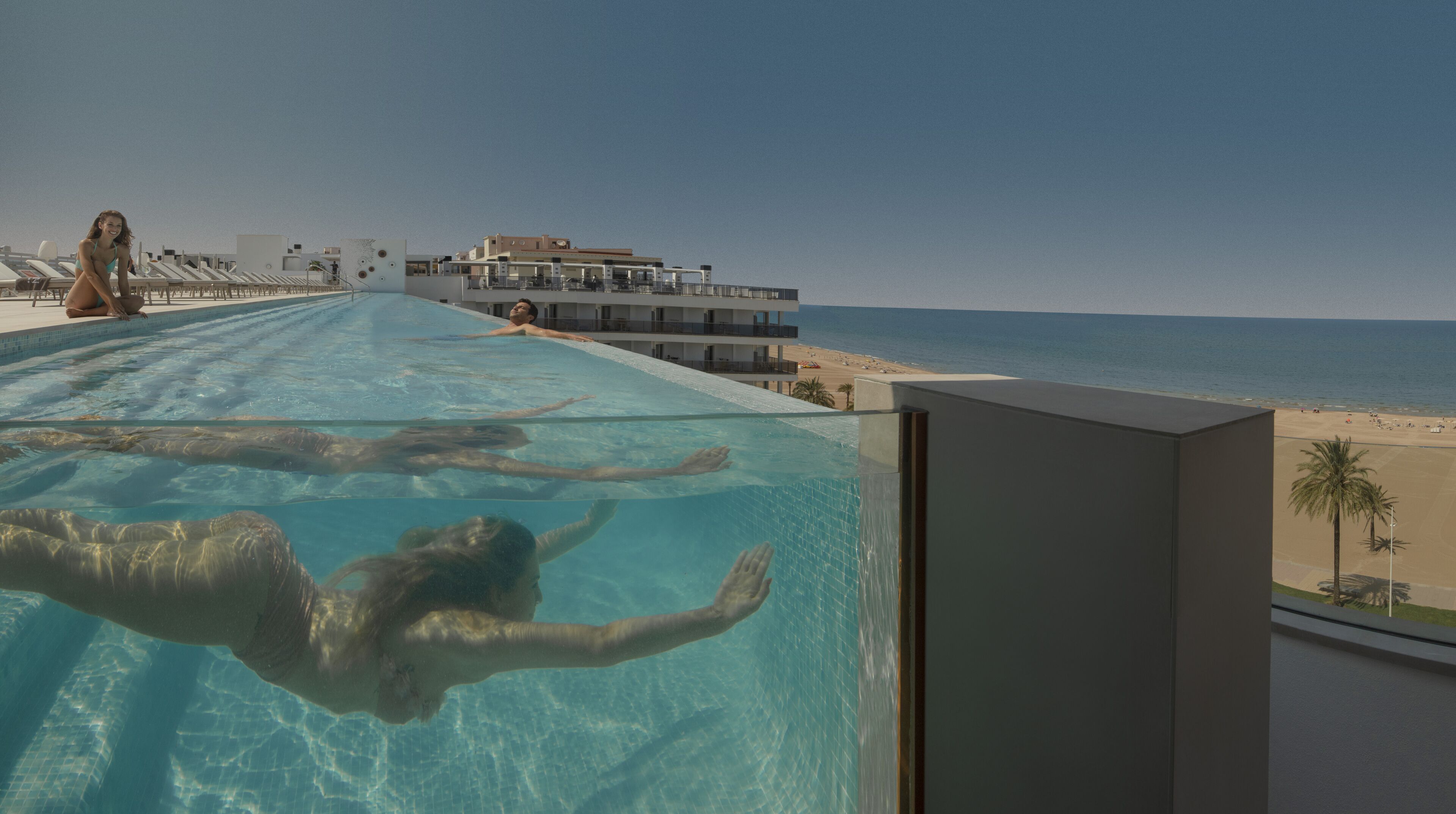 A swimming pool with clear water, a beach view, and a modern building in the background.