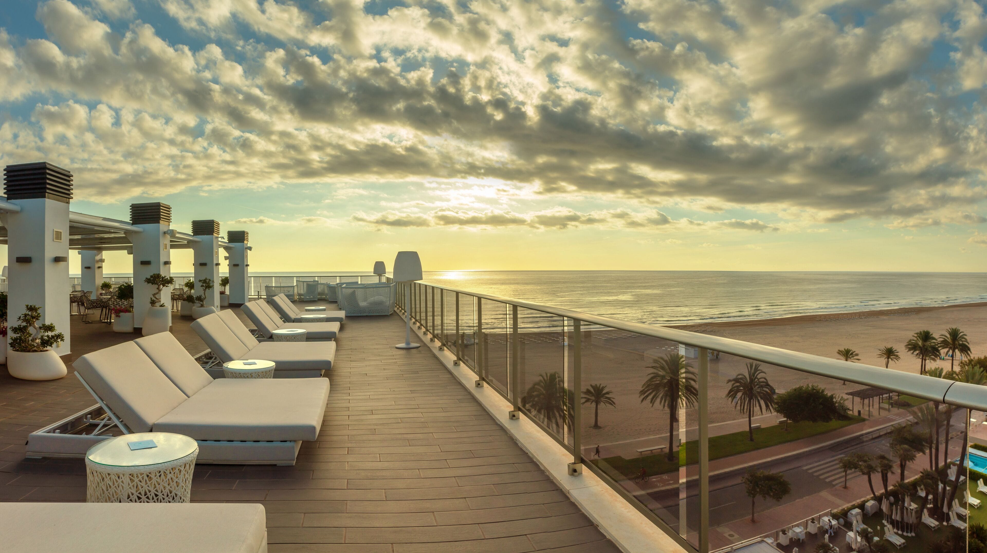 A rooftop terrace with sun loungers, a glass balustrade, and a view of the beach and ocean.