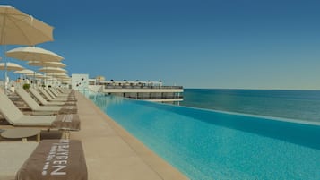 Un área junto a la piscina con sillas de descanso y sombrillas, una piscina azul cristalina y vista al océano.