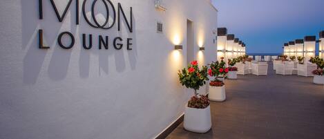 A Mon Lounge with a white exterior wall, outdoor seating area with white chairs, and potted plants with red flowers.