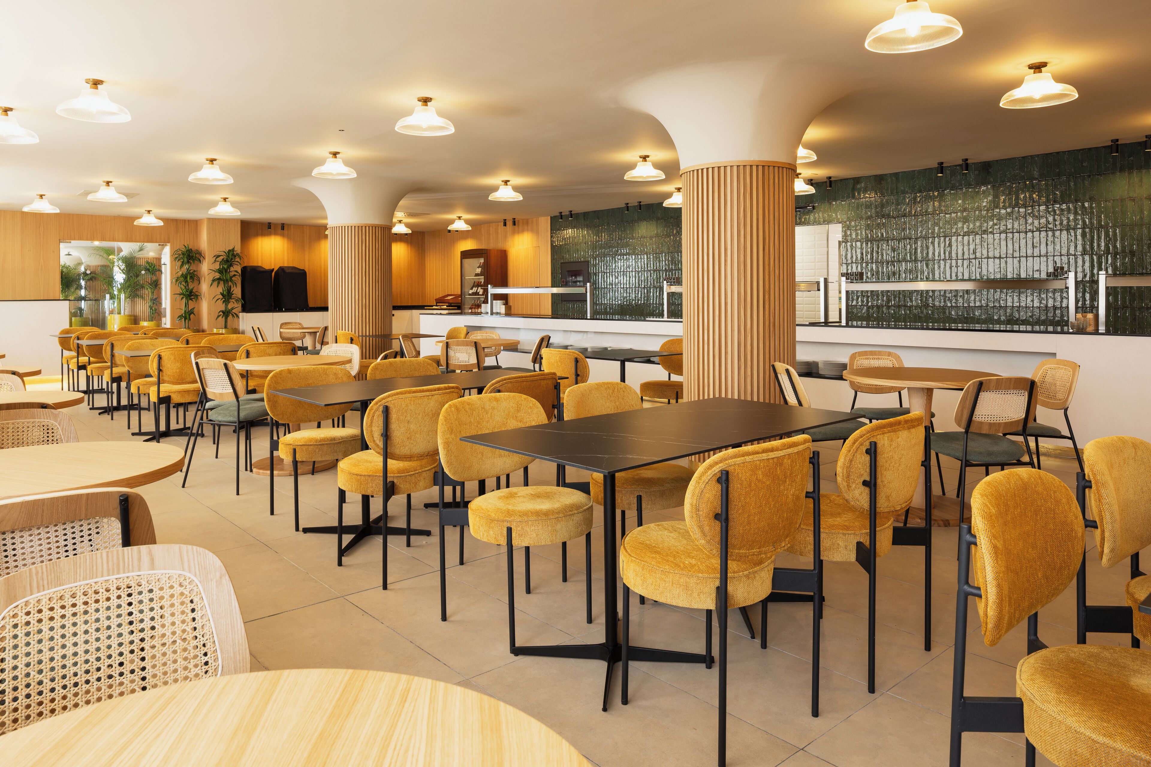 A modern restaurant with yellow chairs and tables, a bar area, and a view of the city through large windows.