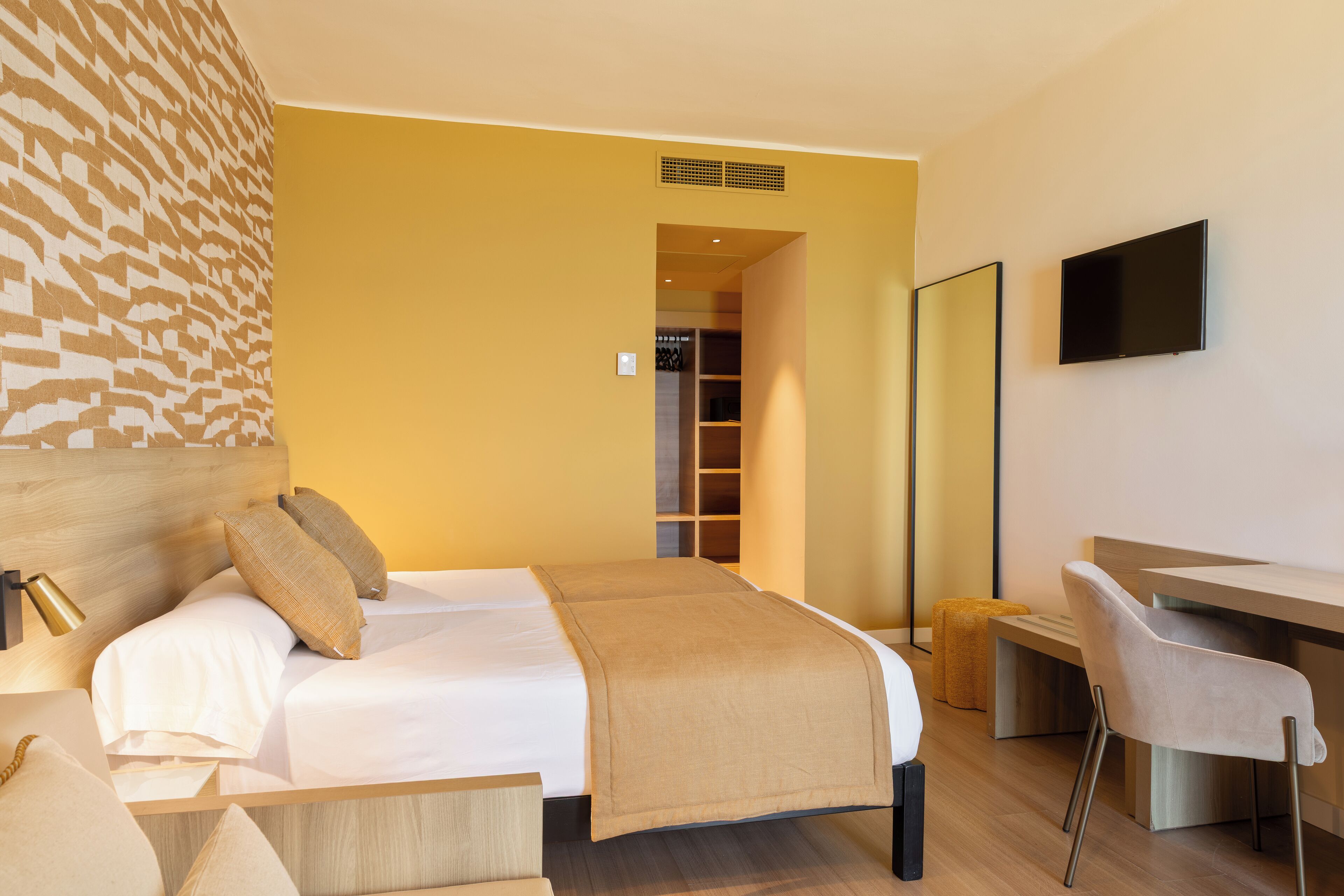 A hotel room with a bed, bedside table, chair, desk, and a TV mounted on the wall.