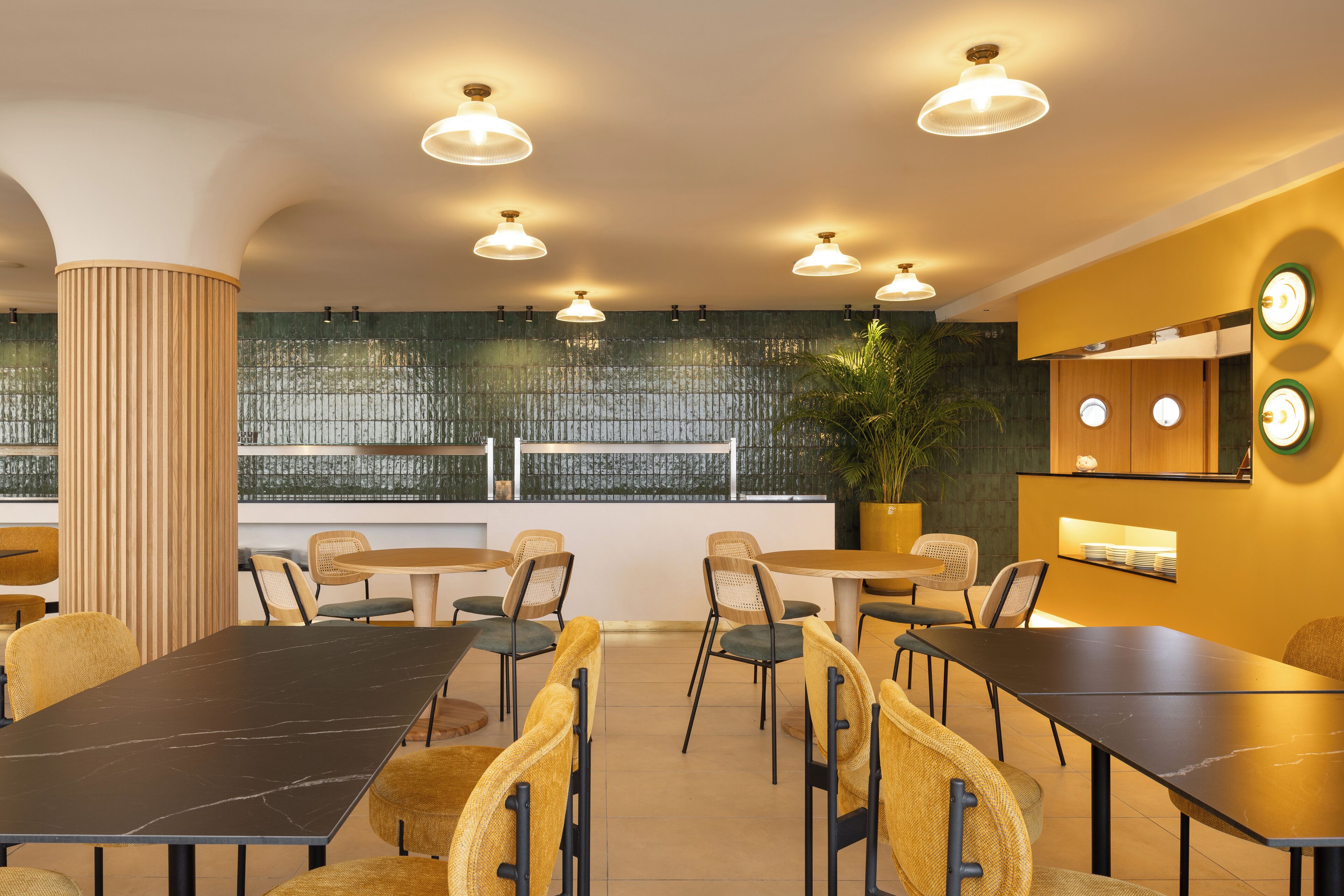 A modern dining area with multiple tables and chairs, a large potted plant, and a view through a patterned glass wall.