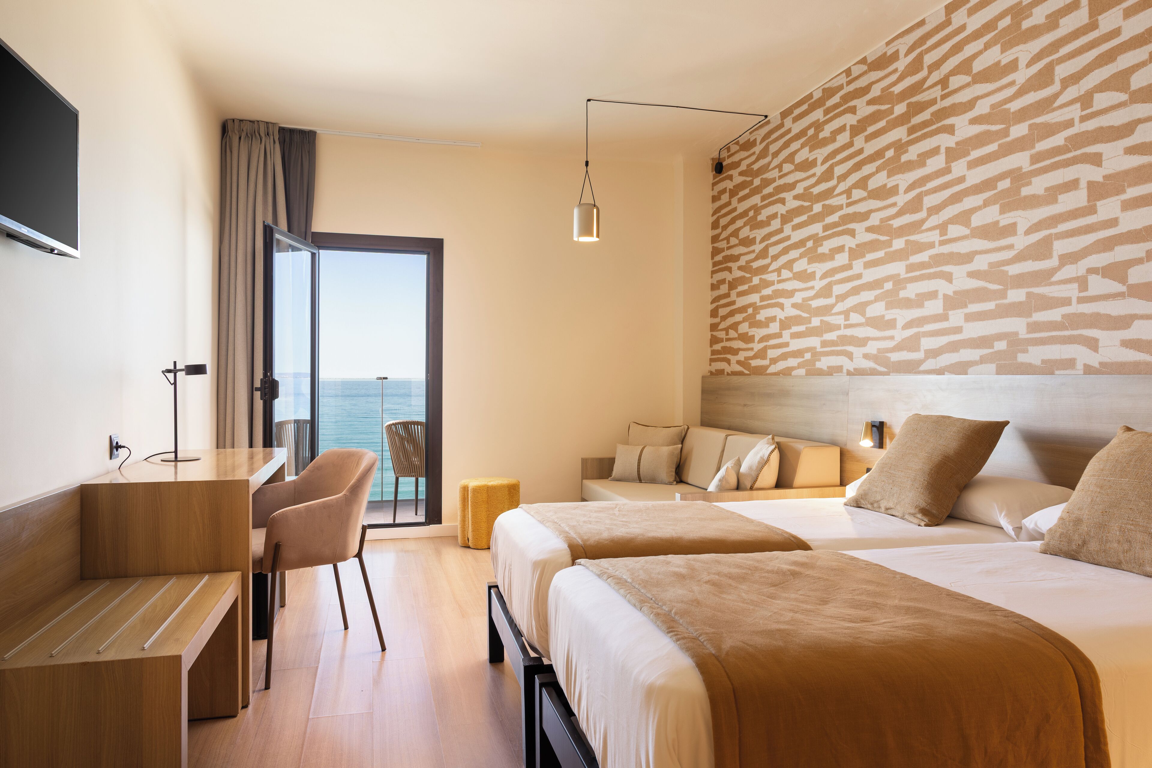 Ein Hotelzimmer mit zwei Betten, einem Schreibtisch und einem Stuhl. Ein großes Fenster bietet Blick aufs Meer.