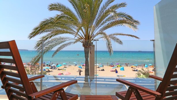 Vista a la playa desde un balcón con sillas de madera, una palmera y cielo azul despejado.
