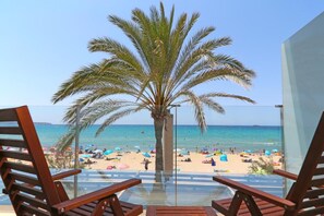 A beach view from a balcony with wooden chairs, a palm tree, and a clear blue sky. - Hotel Eden Palma Playa (Palma de Mallorca)