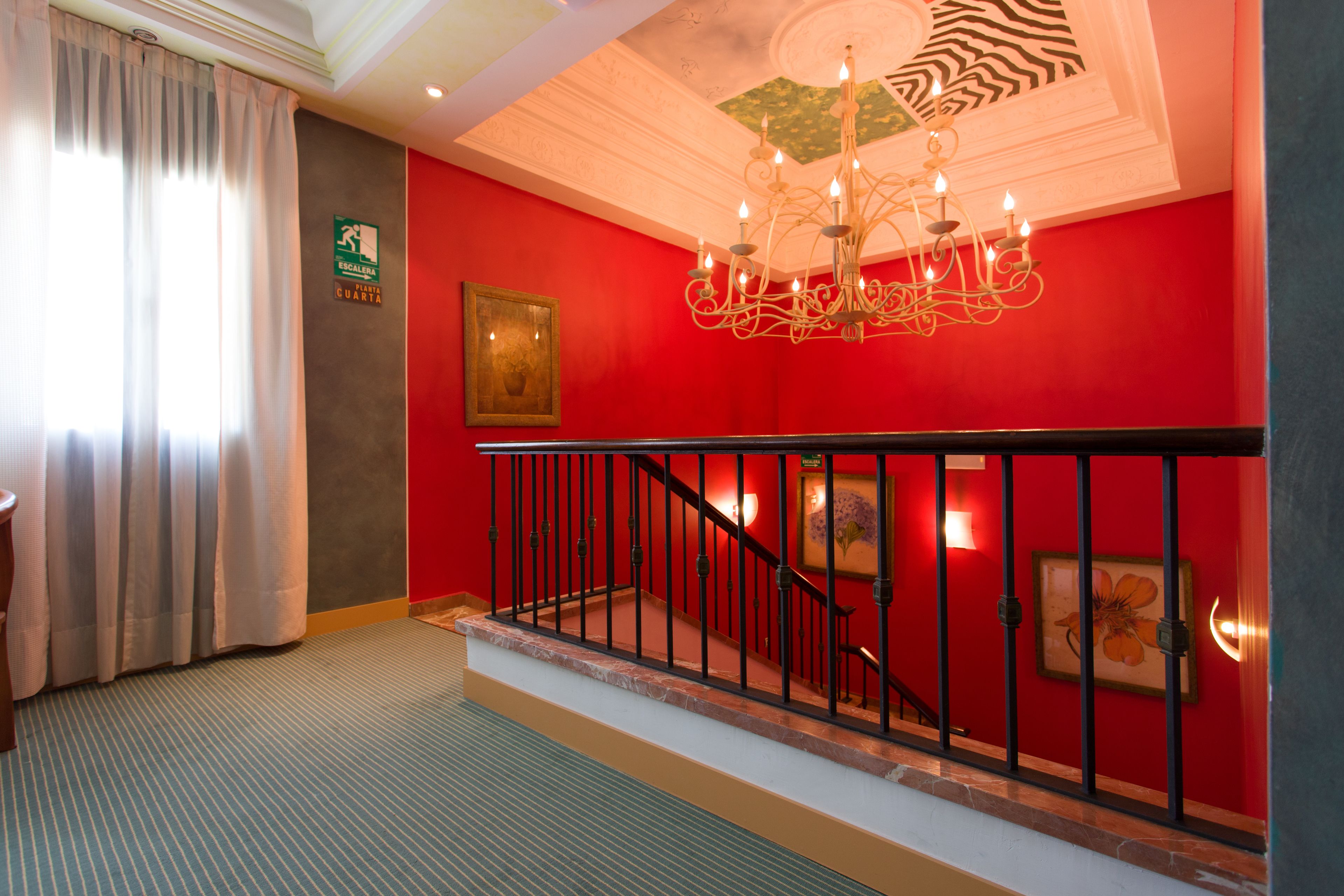 A hotel lobby with a red wall, a chandelier, a staircase with black railings, and a sign indicating the location.