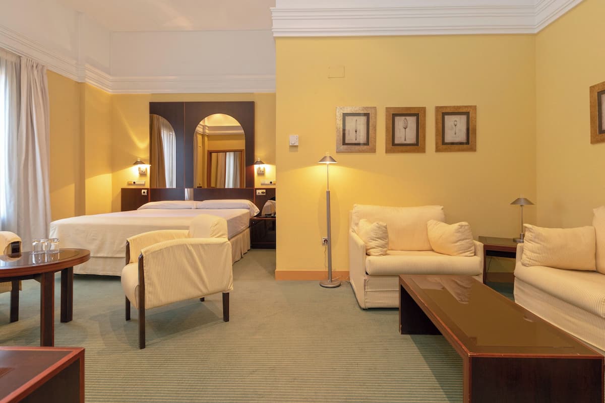 Une chambre d’hôtel avec deux lits simples, un coin salon avec un canapé et des fauteuils, une petite table et une lampe sur pied.