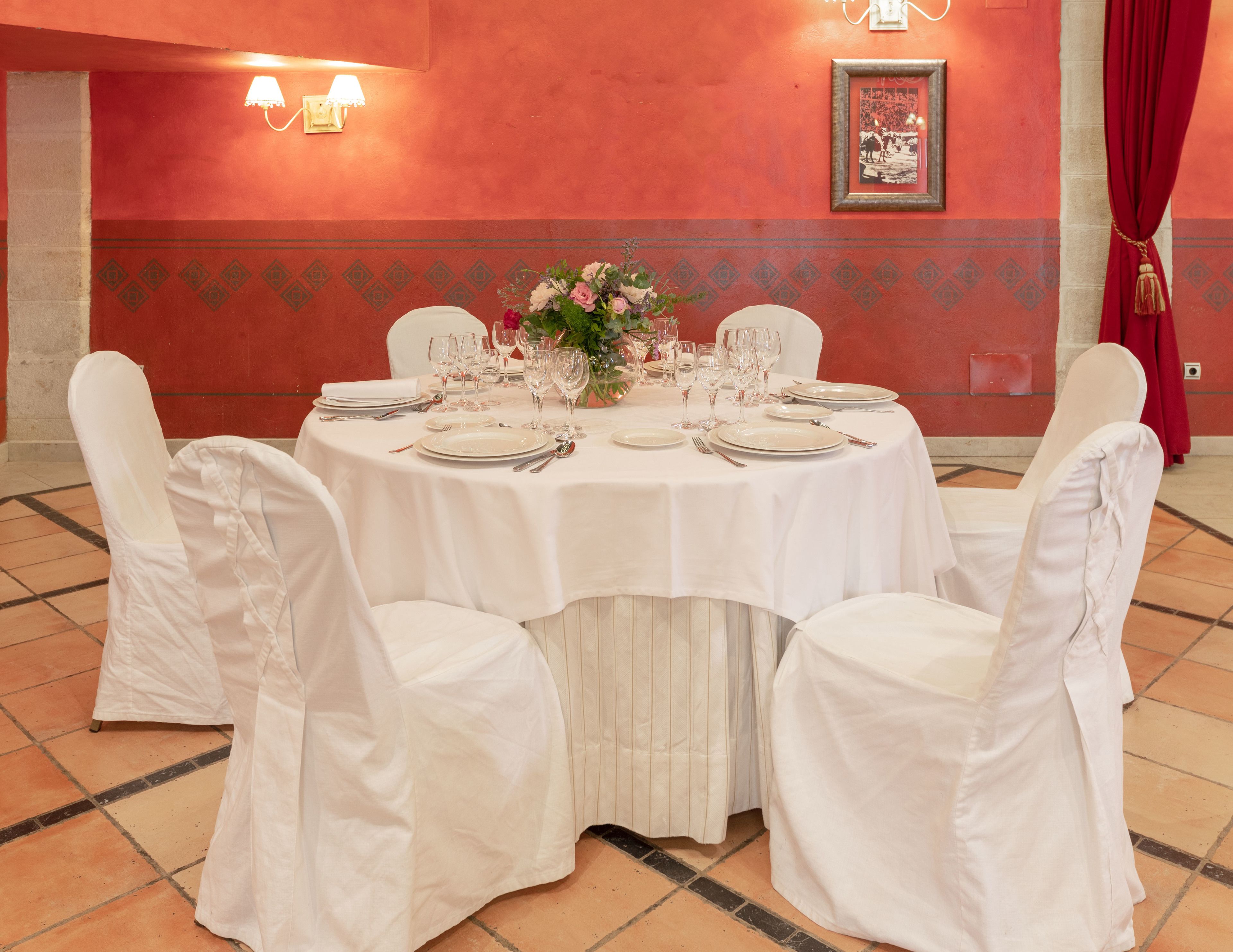 Une table de salle à manger ronde, recouverte d’une nappe blanche, entourée de chaises, dressée avec de la vaisselle et des verres, dans une pièce aux murs rouges et au sol carrelé.