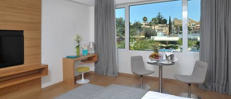 A modern hotel room with a large bed, a TV mounted on the wall, a small desk with a chair, and a window offering a city view.