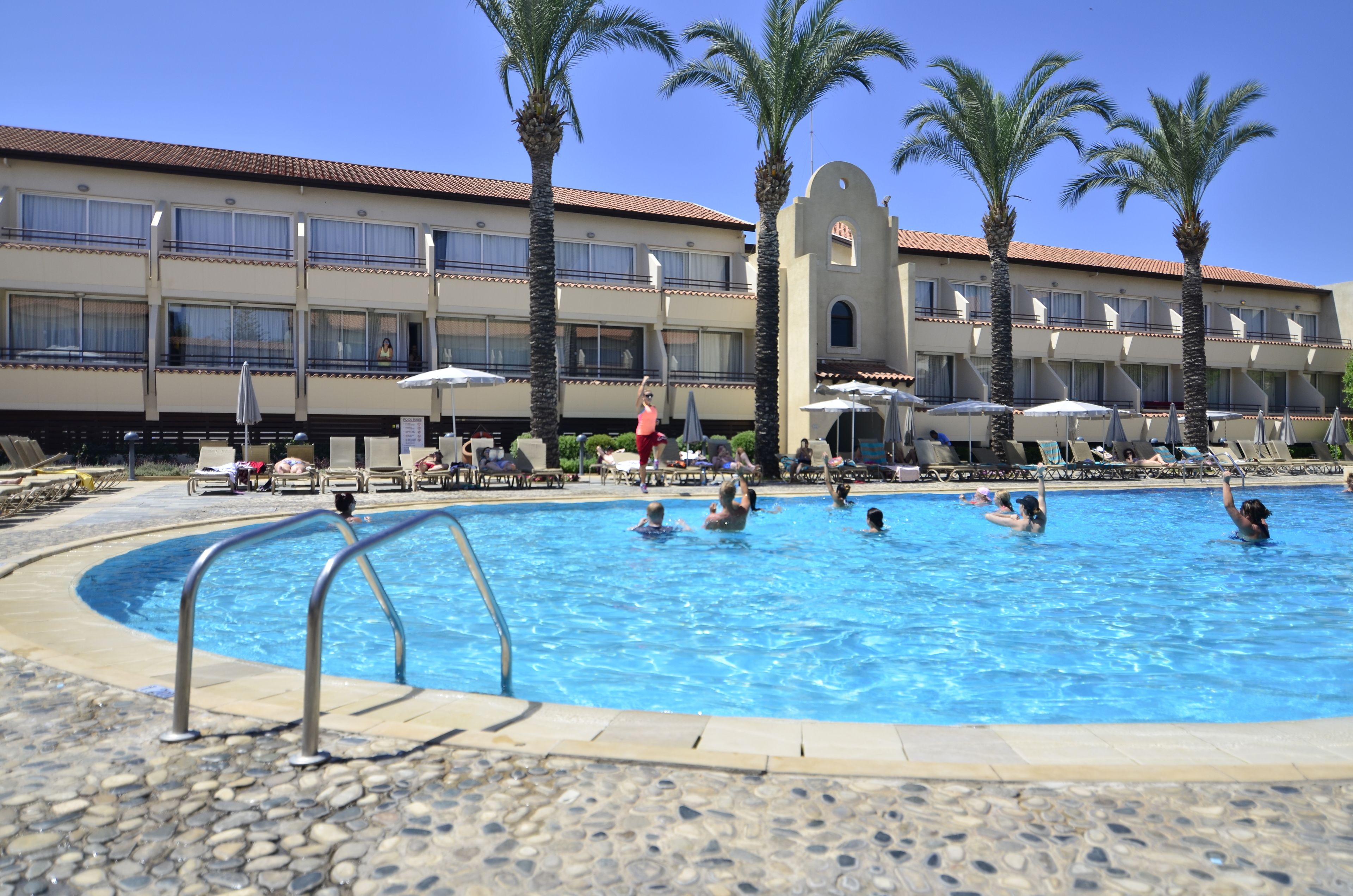 A swimming pool with lounge chairs and umbrellas, surrounded by palm trees and a hotel building with balconies.
