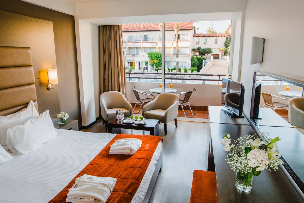 A hotel room with a bed, a desk with a TV, a balcony with chairs, and a view of buildings.